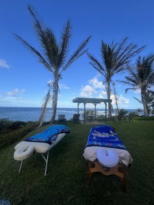 Two massage tables set on the grass overlooking the ocean, framed by palm trees on Kauaʻi